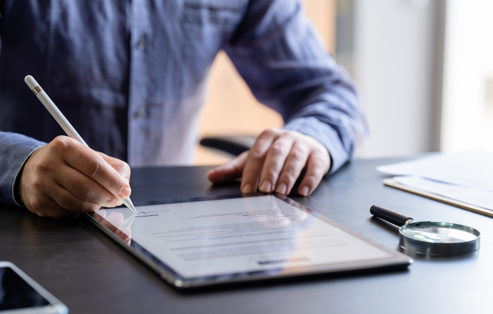 Close-up of a hand signing a contract on a tablet, with a magnifying glass reviewing the terms