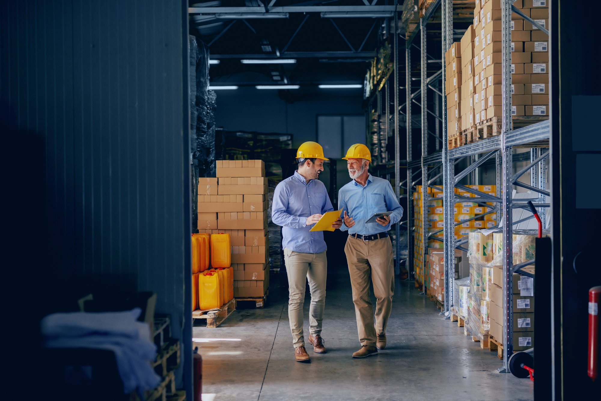 Two supply chain professionals in hard hats walking a warehouse floor together, reviewing a supplier scorecard